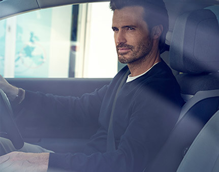 Ein Mann sitzt in einem Auto auf dem Fahrersitz und schaut aus dem Fenster. Er trägt einen dunkelblauen Pullover.