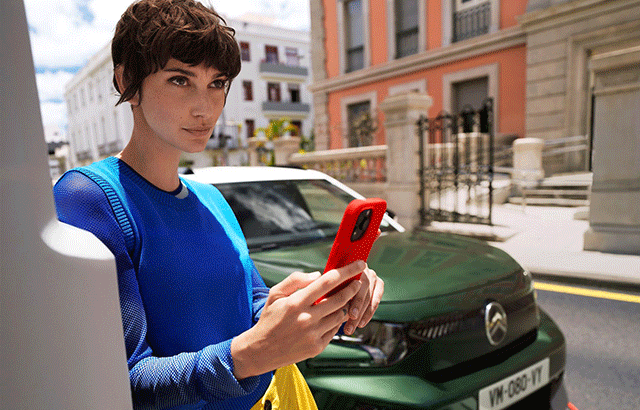 Eine Person mit kurzem braunem Haar trägt einen blauen Pullover und hält ein rotes Smartphone in der Hand. Sie steht vor einem grünen Auto, das auf einer städtischen Straße geparkt ist, mit Gebäuden im Hintergrund.